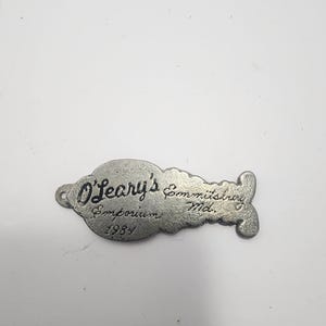 May include: A vintage, silver-toned metal tag from O'Leary's Emporium, Emmitsburg, Md. The tag has a unique shape with a hole on one end and a decorative flourish on the other. The text "O'Leary's Emporium 1984" is engraved on the tag.