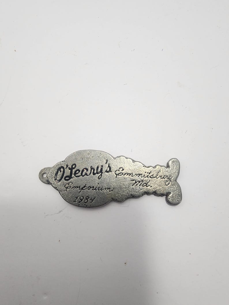 May include: A vintage, silver-toned metal tag from O'Leary's Emporium, Emmitsburg, Md. The tag has a unique shape with a hole on one end and a decorative flourish on the other. The text "O'Leary's Emporium 1984" is engraved on the tag.