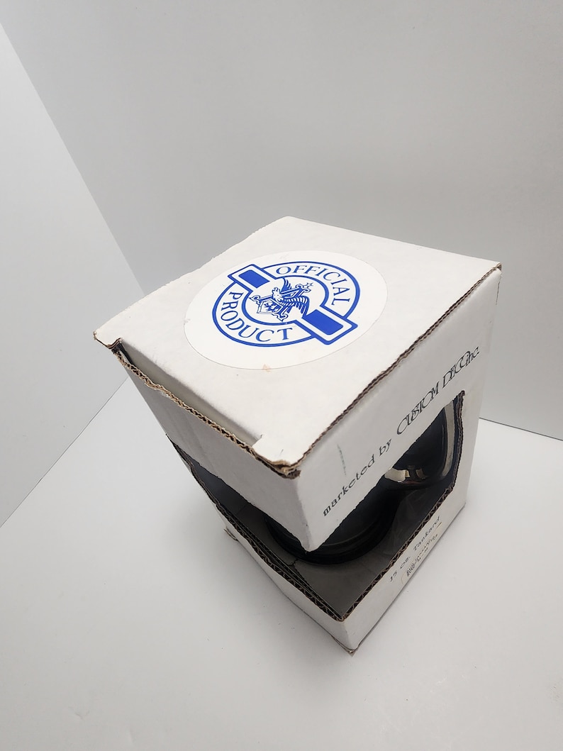 Puede incluir: Una caja blanca con una pegatina azul y blanca que dice "Producto Oficial" y un art&iacute;culo negro y plateado en su interior. La caja tambi&eacute;n dice "comercializado por Custom Decals".