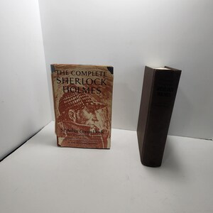 May include: A hardcover book titled The Complete Sherlock Holmes by Sir Arthur Conan Doyle with a tan dust jacket featuring a brown illustration of a detective in a deerstalker hat, positioned next to a plain dark brown book spine standing upright.
