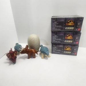 Könnte beinhalten: Ein Set mit drei Jurassic World Minis Blindboxen, jede mit einem anderen Dinosaurier. Die Boxen sind übereinander gestapelt und haben ein schwarz-graues Design mit dem Jurassic World Logo und dem Schriftzug "Minis". Es gibt auch vier Dinosaurierspielzeuge, darunter einen roten Dinosaurier, einen braunen Dinosaurier, einen blauen Dinosaurier und ein weißes Dinosauriereier.