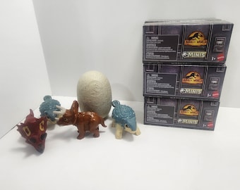 May include: A set of three Jurassic World Minis blind boxes, each featuring a different dinosaur. The boxes are stacked on top of each other and have a black and gray design with the Jurassic World logo and the words "Minis" printed on them. There are also four dinosaur toys, including a red dinosaur, a brown dinosaur, a blue dinosaur, and a white dinosaur egg.