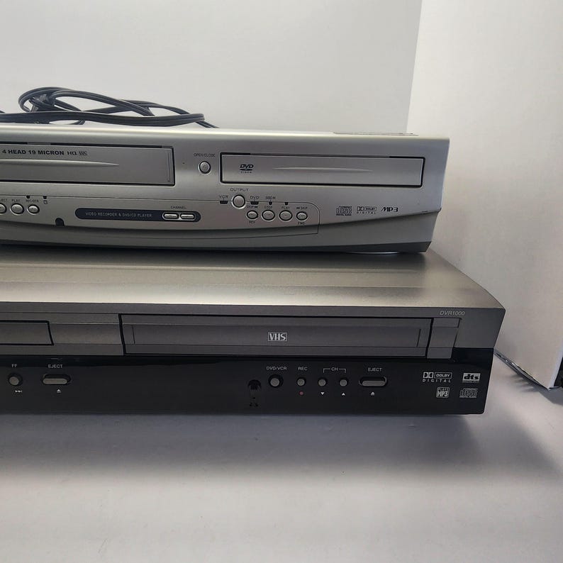 Może przedstawiać: Dwa srebrno-czarne odtwarzacze combo DVD/VHS ułożone jeden na drugim. G&oacute;rny odtwarzacz ma odtwarzacz DVD i odtwarzacz VHS. Dolny odtwarzacz to odtwarzacz DVR/VHS. Oba urządzenia mają r&oacute;żne przyciski i porty.