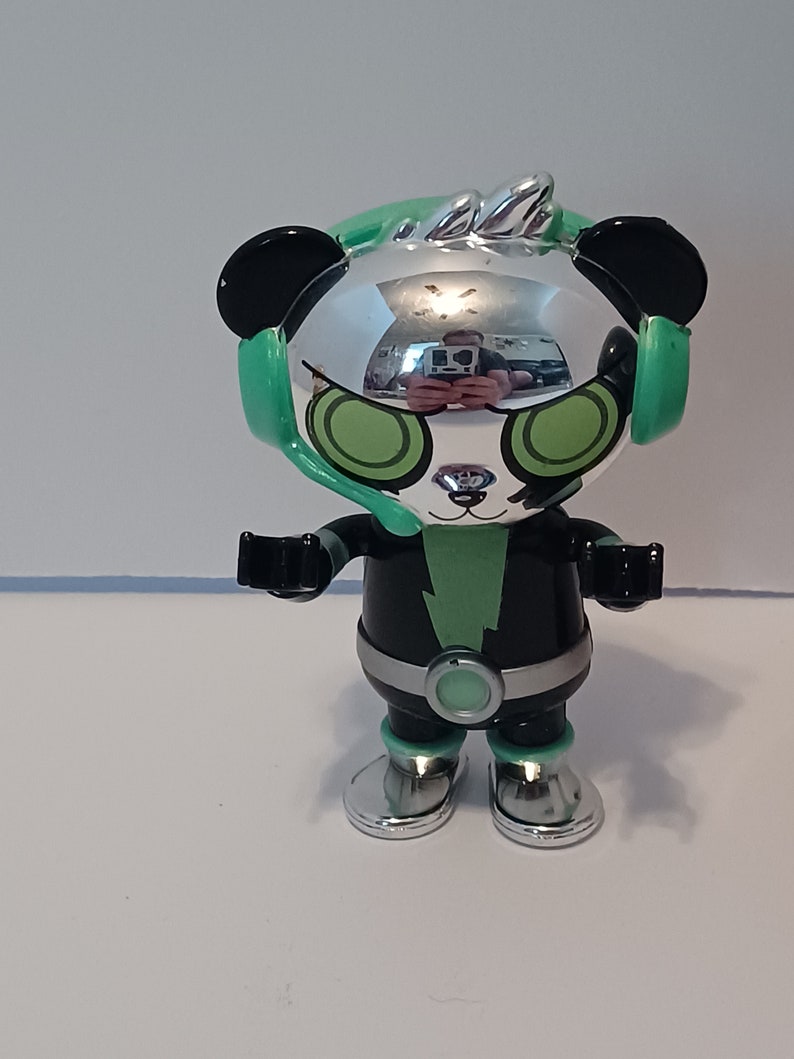 Puede incluir: Un panda de juguete plateado y negro que lleva unos auriculares verdes con un rayo verde en el pecho. El panda tiene ojos verdes y un mohawk plateado.