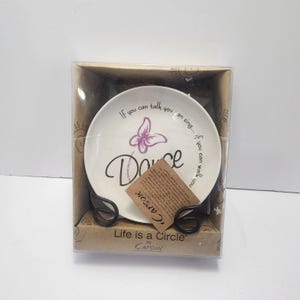 Puede incluir: Un plato decorativo blanco con la palabra "Dance" en cursiva, una mariposa morada y el texto "If you can talk you can sing... if you can walk you". El plato está sobre un soporte de metal negro y en una caja transparente.