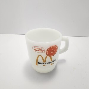 Puede incluir: Taza de McDonald's de cer&aacute;mica blanca con asa. La taza presenta el logotipo de McDonald's en dorado y las palabras "Good Morning" en un bocadillo, junto con un sol sonriente naranja.