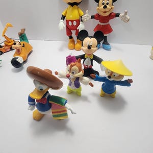Op de afbeelding: Een verzameling kleurrijke Disney-personagefiguren, waaronder Mickey Mouse, Minnie Mouse, Donald Duck, Goofy en Chip en Dale. Mickey Mouse draagt een rode broek en gele schoenen. Donald Duck draagt een blauw matrozenpak en een sombrero.