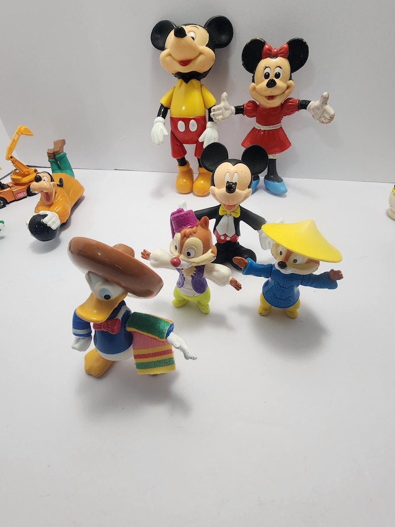 Op de afbeelding: Een verzameling kleurrijke Disney-personagefiguren, waaronder Mickey Mouse, Minnie Mouse, Donald Duck, Goofy en Chip en Dale. Mickey Mouse draagt een rode broek en gele schoenen. Donald Duck draagt een blauw matrozenpak en een sombrero.