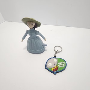 May include: A light blue figurine of a woman wearing a hat and dress, and a keychain with a cartoon character. The keychain has the text "Victory shall be mine!" on a green and blue background.