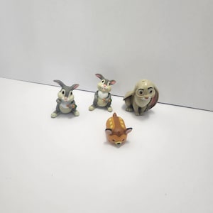 May include: Four small figurines of characters from the Disney movie Bambi. The figurines include three rabbits in various poses, and a small deer figurine. The figurines are in shades of gray, brown, and tan.