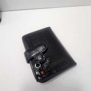 May include: Black wallet with a crocodile skin texture. The wallet has a flap closure with a decorative charm and a zipper pocket. The wallet is made of a shiny material.