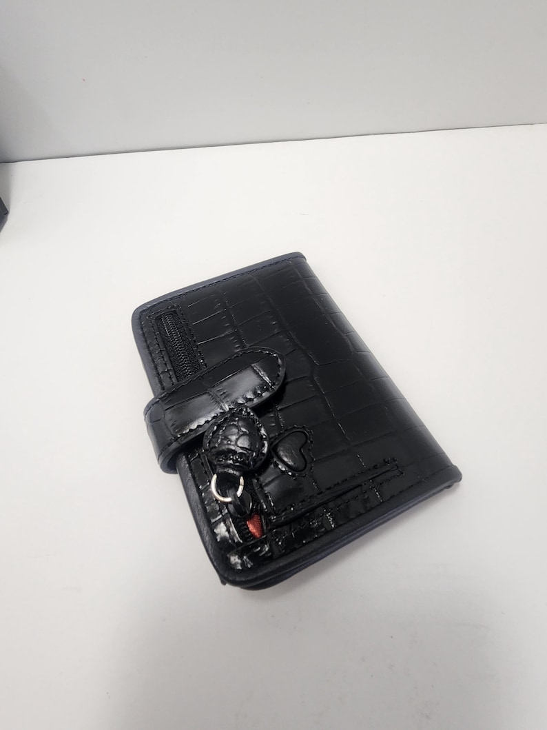 May include: Black wallet with a crocodile skin texture. The wallet has a flap closure with a decorative charm and a zipper pocket. The wallet is made of a shiny material.