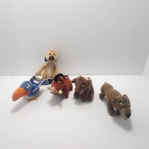May include: A collection of plush toys featuring characters from the animated film "The Lion King." The toys include a tan meerkat, a blue bird with an orange beak, a brown warthog, a brown lion cub, and a tan lioness.