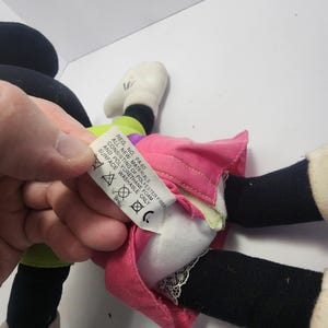 May include: A Minnie Mouse plush toy, featuring a pink skirt, black legs, white gloves, and a green and purple top. The toy's tag reads "REG. NO. PA-60 ALL NEW MATERIALS CONSISTING OF POLYESTER FIBER AND POLYURETHANE FOAM SURFACE WASHABLE ONLY."