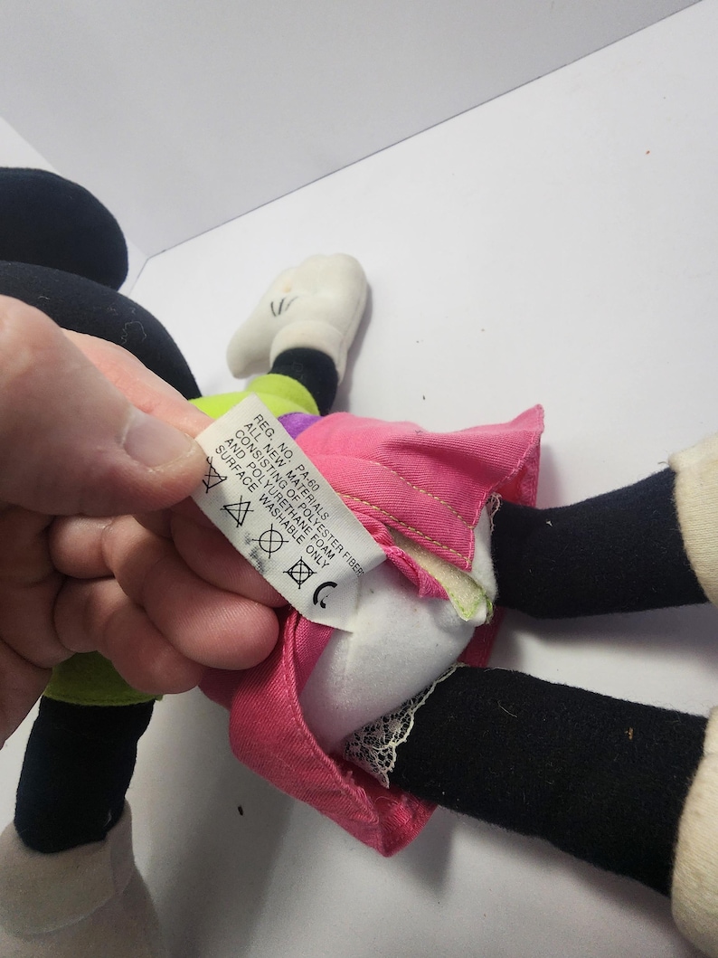 May include: A Minnie Mouse plush toy, featuring a pink skirt, black legs, white gloves, and a green and purple top. The toy's tag reads "REG. NO. PA-60 ALL NEW MATERIALS CONSISTING OF POLYESTER FIBER AND POLYURETHANE FOAM SURFACE WASHABLE ONLY."