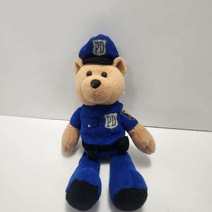 Op de afbeelding: Een pluchen teddybeer gekleed in een blauw politie-uniform, inclusief een hoed en een shirt met "PD" patches. De beer heeft bruine vacht op zijn hoofd, armen en benen, met zwarte poten en een zwarte riem.
