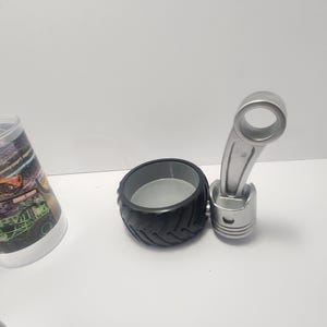 May include: A black tire-shaped container with a gray interior, paired with a silver piston-shaped object. The objects are on a white surface. The items appear to be decorative or novelty items.