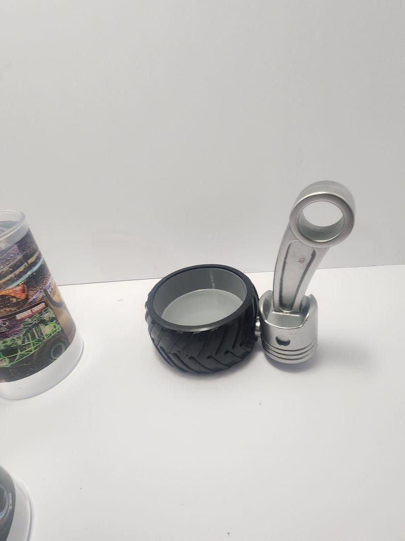 May include: A black tire-shaped container with a gray interior, paired with a silver piston-shaped object. The objects are on a white surface. The items appear to be decorative or novelty items.