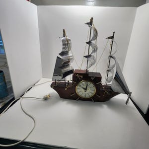 May include: A ship-shaped electric clock featuring a dark brown wooden hull and three masts with reflective silver metal sails. The clock face is circular with black numerals and hands, integrated into the center of the ship's hull with a white power cord.