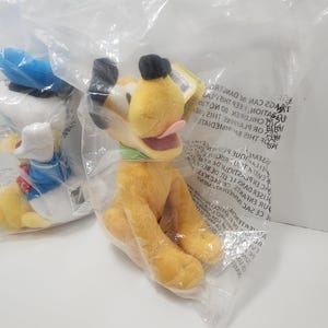 Pu&ograve; includere: Un peluche giallo di Pluto con orecchie nere, un colletto verde e una lingua rosa, confezionato in un sacchetto di plastica trasparente. Un altro peluche &egrave; parzialmente visibile a sinistra.