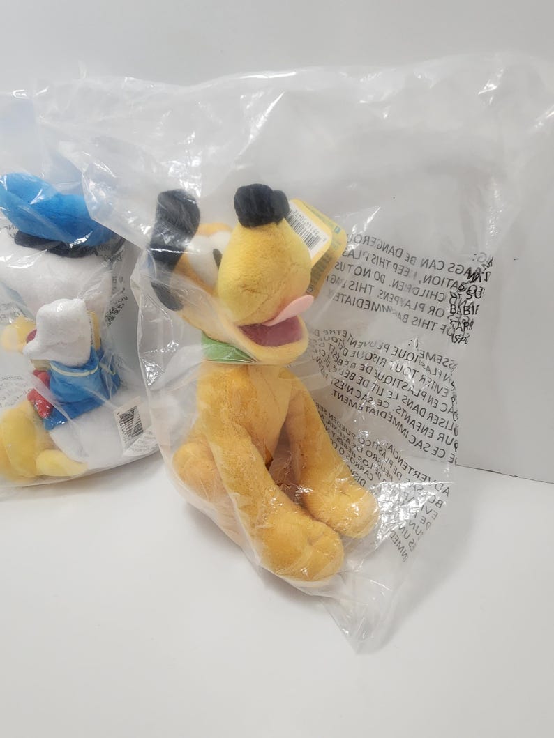 Pu&ograve; includere: Un peluche giallo di Pluto con orecchie nere, un colletto verde e una lingua rosa, confezionato in un sacchetto di plastica trasparente. Un altro peluche &egrave; parzialmente visibile a sinistra.
