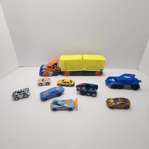 May include: A collection of toy cars in various colors, including blue, yellow, orange, and gold. The assortment features a large yellow and orange semi-truck and several smaller cars with unique designs. The toys are arranged on a white surface.