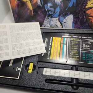 Peut inclure: Un plateau de jeu en plastique noir avec un curseur jaune, une r&egrave;gle et un tableau avec le texte "OFFENSE PLAY SELECTOR" et "DEFENSE SELECTOR". Le plateau est pour un jeu de football.