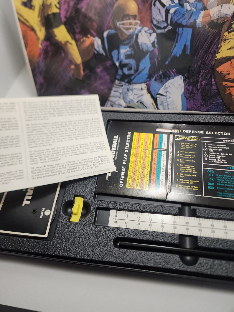 Peut inclure: Un plateau de jeu en plastique noir avec un curseur jaune, une r&egrave;gle et un tableau avec le texte "OFFENSE PLAY SELECTOR" et "DEFENSE SELECTOR". Le plateau est pour un jeu de football.