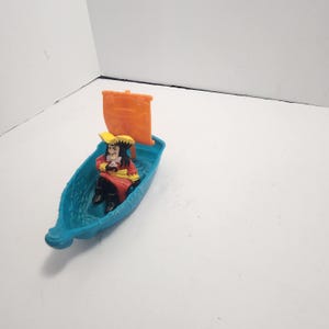May include: A toy boat with a Captain Hook figurine. The boat is turquoise with an orange sail. Captain Hook is wearing a red coat, black pants, and a yellow hat. The figurine is in a seated position.