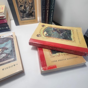 May include: A collection of books from "A Series of Unfortunate Events" by Lemony Snicket. The books have colorful covers with illustrations and titles like "The Vile Village" and "The Ersatz Elevator". The books are stacked and arranged on a white surface.