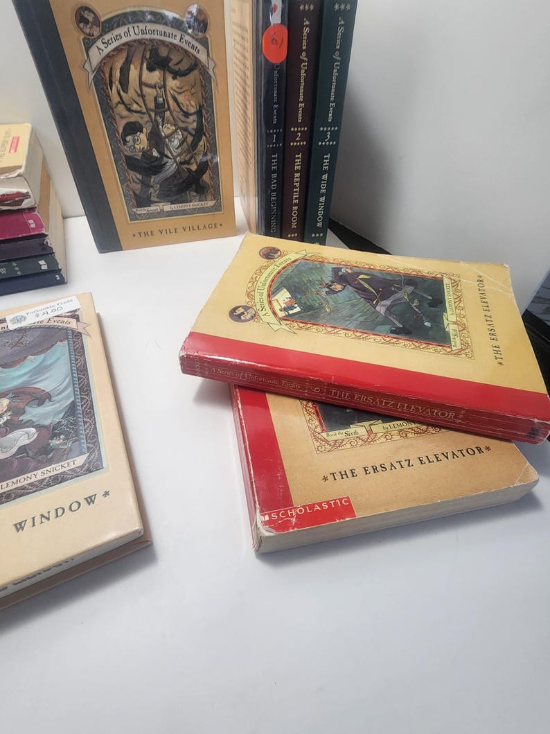 May include: A collection of books from "A Series of Unfortunate Events" by Lemony Snicket. The books have colorful covers with illustrations and titles like "The Vile Village" and "The Ersatz Elevator". The books are stacked and arranged on a white surface.