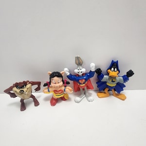 May include: Set of four plastic cartoon character figurines. The characters are Bugs Bunny as Superman, Daffy Duck as Batman, Tweety Bird as Wonder Woman, and Taz as a brown and black devil.