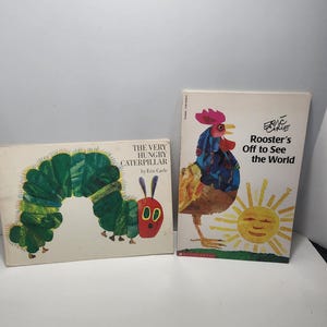 Puede incluir: Dos libros infantiles de Eric Carle. La Oruga Muy Hambrienta presenta una oruga verde con ojos rojos y una boca roja. El Gallo que Sale a Ver el Mundo presenta un gallo con una cresta roja y un sol amarillo.