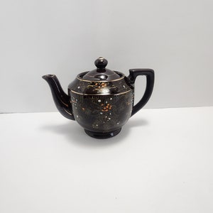 May include: A small brown teapot with a floral design in gold and orange. The teapot has a lid and a spout.