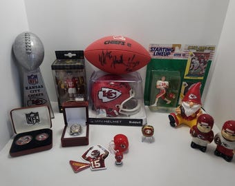 May include: A collection of Kansas City Chiefs memorabilia, including a football, mini helmet, figurines, a ring, and a trophy. The items are red, white, and gold, with the Chiefs logo and player numbers visible. The football has signatures.