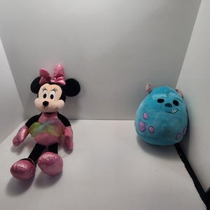May include: Two plush toys: Minnie Mouse and Sulley from Monsters, Inc. Minnie Mouse has a pink bow, dress, and shoes. Sulley is a round, blue monster with purple spots. Both toys are on a white background.