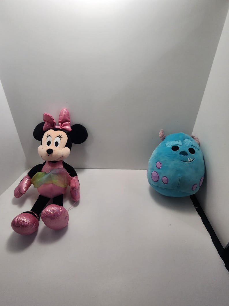 May include: Two plush toys: Minnie Mouse and Sulley from Monsters, Inc. Minnie Mouse has a pink bow, dress, and shoes. Sulley is a round, blue monster with purple spots. Both toys are on a white background.