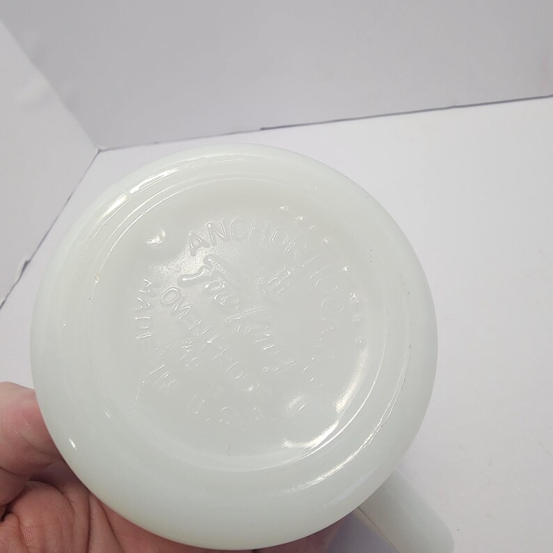Puede incluir: Una taza blanca y opaca con base redondeada y asa. La base de la taza tiene el texto en relieve "Anchor Hocking" y "Made in U.S.A.". La taza est&aacute; limpia y parece estar en buen estado.