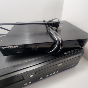 K&ouml;nnte beinhalten: Ein schwarzer Samsung Blu-ray Disc-Player auf einem schwarzen Magnavox DVD/VHS-Kombi-Player. Beide Player haben einen silbernen Rand um die Vorderseite.