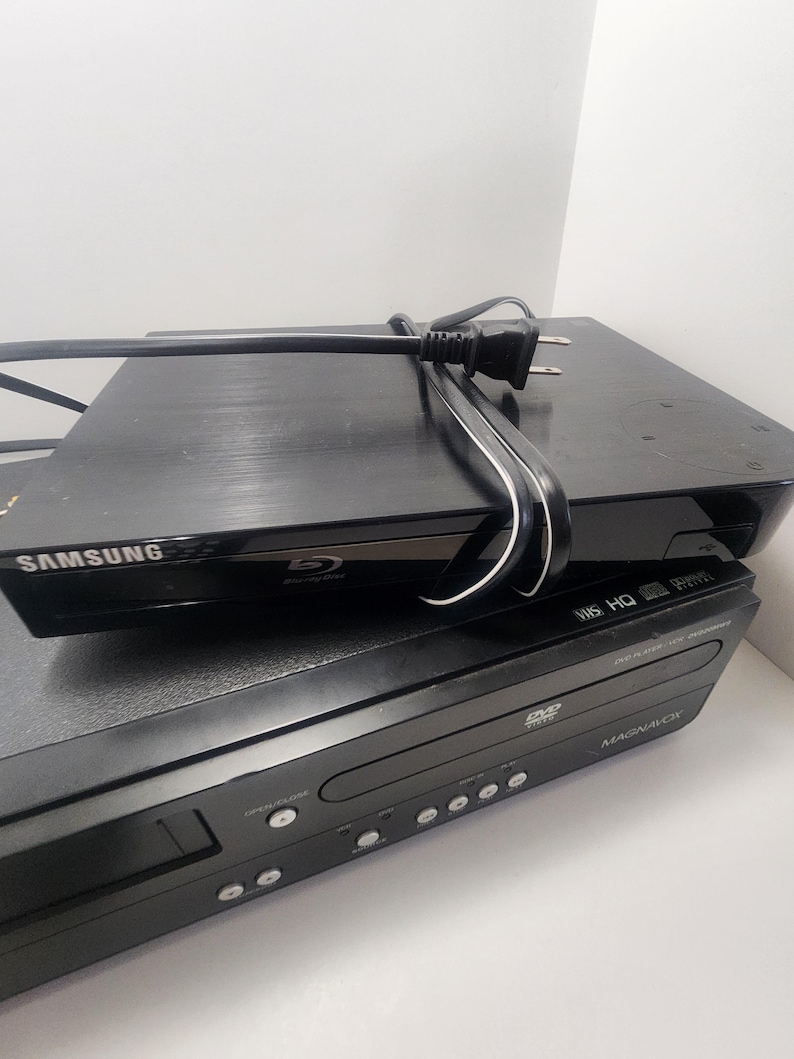 K&ouml;nnte beinhalten: Ein schwarzer Samsung Blu-ray Disc-Player auf einem schwarzen Magnavox DVD/VHS-Kombi-Player. Beide Player haben einen silbernen Rand um die Vorderseite.