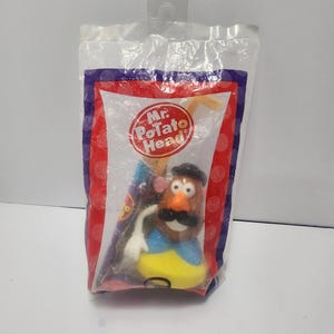 May include: A new, unopened Mr. Potato Head toy in its original packaging. The packaging is a clear plastic bag with a red, white, and blue border. The Mr. Potato Head toy has a brown body, an orange nose, and a black mustache.