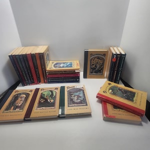 May include: A collection of hardcover books with colorful illustrations. The books have various titles and are arranged in stacks and rows. The covers feature detailed artwork and text, with a mix of red, tan, and dark-colored spines. The books are on a white surface.