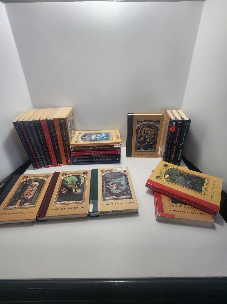May include: A collection of hardcover books with colorful illustrations. The books have various titles and are arranged in stacks and rows. The covers feature detailed artwork and text, with a mix of red, tan, and dark-colored spines. The books are on a white surface.