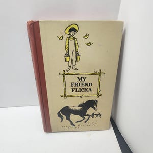 May include: A vintage hardcover book titled "My Friend Flicka." The cover displays a black illustration of a horse and foal, a boy with buckets, and the title in a rustic frame. The book has a tan cover with a red spine.