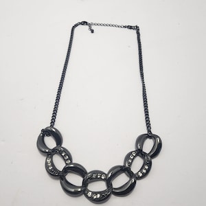 Può includere: Una collana grigio scuro con catena e grandi maglie ovali. Alcune maglie sono impreziosite da piccoli cristalli trasparenti. La collana è esposta su sfondo bianco.