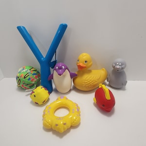 May include: A set of bath toys including a yellow rubber duck, a purple penguin, a gray seal, a red fish, a yellow and green ball, a yellow ring, and a blue slingshot.