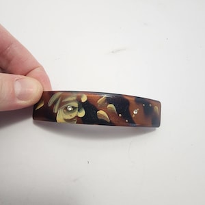 May include: A brown and black hair barrette with a tortoiseshell pattern. The barrette features a painted floral design with cream and brown accents and two small, clear rhinestones.