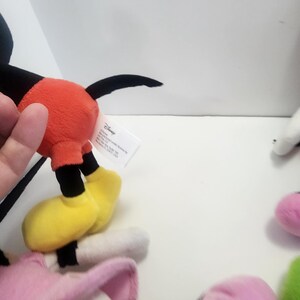 May include: A set of four plush Disney characters. Mickey Mouse is wearing his signature red shorts and yellow shoes. Minnie Mouse is wearing a pink dress and white shoes. Donald Duck is wearing a blue sailor shirt and white trousers. Goofy is wearing a purple shirt and yellow trousers. The characters are all smiling and have big eyes.