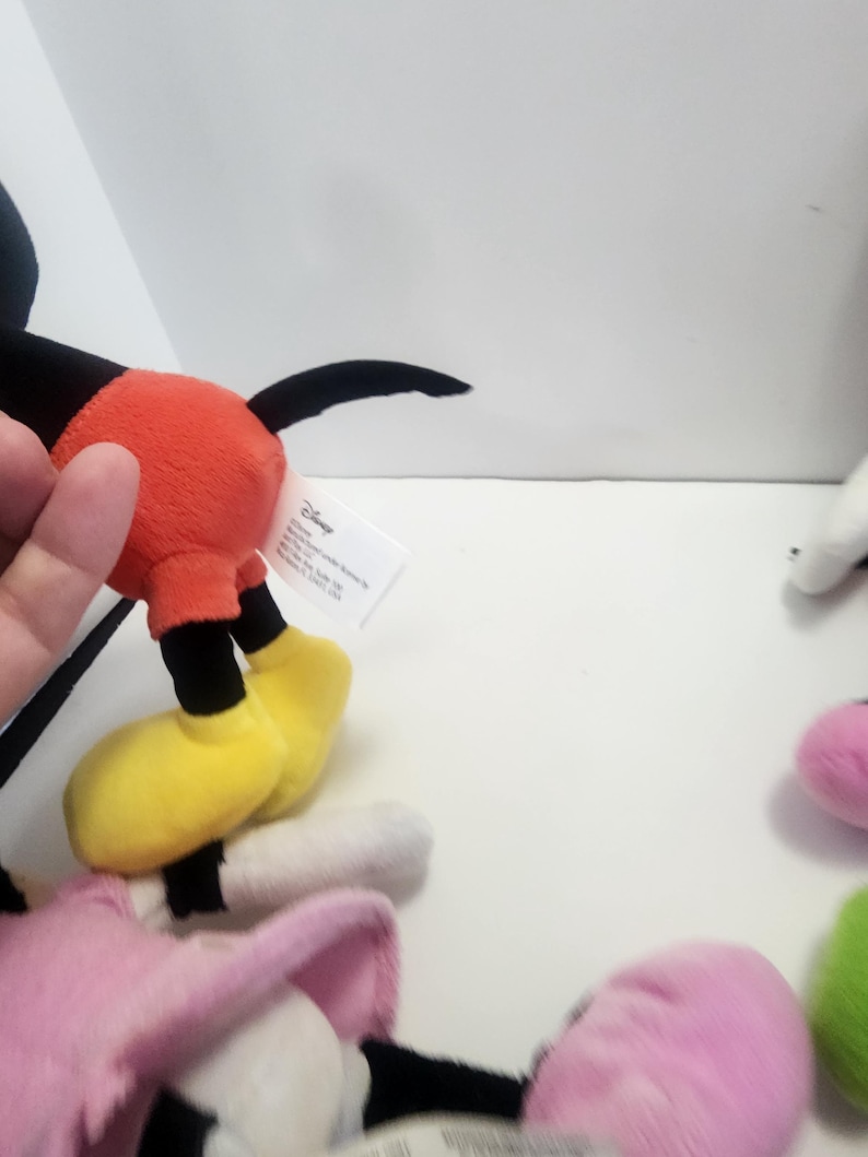 May include: A set of four plush Disney characters. Mickey Mouse is wearing his signature red shorts and yellow shoes. Minnie Mouse is wearing a pink dress and white shoes. Donald Duck is wearing a blue sailor shirt and white trousers. Goofy is wearing a purple shirt and yellow trousers. The characters are all smiling and have big eyes.