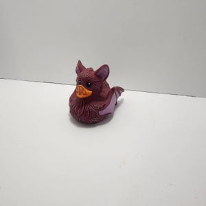 Op de afbeelding: Een rubberen badeend in de vorm van een vleermuis. De vleermuis-eend is voornamelijk diep bordeauxrood van kleur met paarse accenten op de oren en vleugels. Het heeft een oranje snavel, blauwe ogen en een donzige textuur.